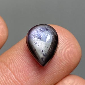May include: A teardrop-shaped gemstone with a dark, glossy surface. The gem has a mix of dark blue, gray, and black hues, with a reflective, polished appearance. The stone is held between two fingers, showcasing its size and shape.