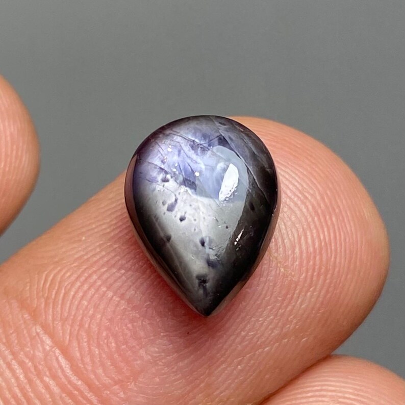 May include: A teardrop-shaped gemstone with a dark, glossy surface. The gem has a mix of dark blue, gray, and black hues, with a reflective, polished appearance. The stone is held between two fingers, showcasing its size and shape.