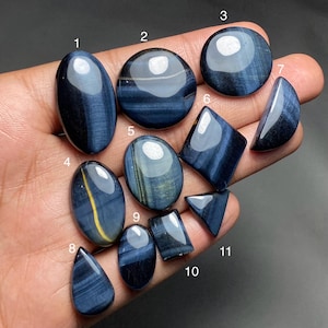 May include: A collection of polished blue tiger's eye gemstones in various shapes, including oval, round, and teardrop. The stones display chatoyancy with bands of color and a golden yellow stripe in some. The gemstones are displayed on a dark background.