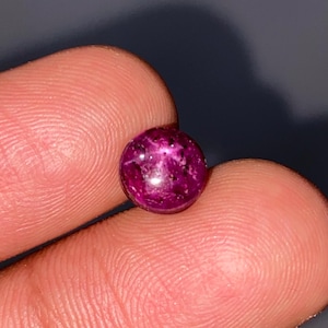 Puede incluir: Una pequeña gema redonda y pulida, de color magenta intenso con inclusiones oscuras. La gema se sostiene entre dos dedos, mostrando su tamaño y textura. La piedra parece ser un cabujón, con una superficie lisa y redondeada.