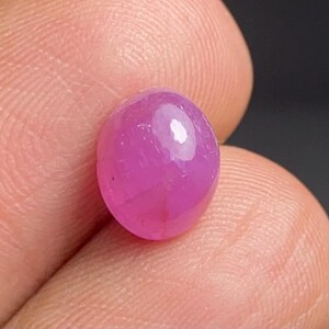 Puede incluir: Una gema rosa pulida, de forma ovalada. La gema tiene una superficie lisa y reflectante con un punto de luz brillante. La piedra se sostiene entre dos dedos, mostrando su tamaño y color.