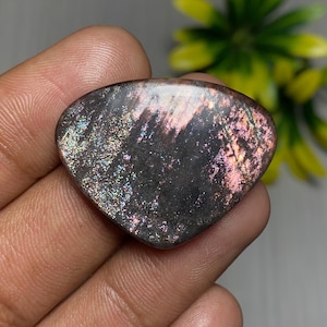 May include: A heart-shaped gemstone with a dark base and shimmering pink and silver inclusions. The polished stone has a smooth, reflective surface. The gemstone is held in a hand. Likely used for jewellery or decorative purposes.