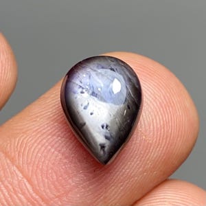 May include: A teardrop-shaped gemstone with a dark, lustrous surface. The gem displays a mix of deep purple and gray hues, with subtle internal patterns and a reflective, polished finish. The stone is held between fingers, showcasing its size and shape.