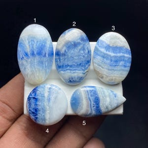 May include: Five polished gemstones with a blue and white banded pattern. The stones are oval, round, and teardrop-shaped, displayed on a white surface. The gemstones have a natural, organic appearance.