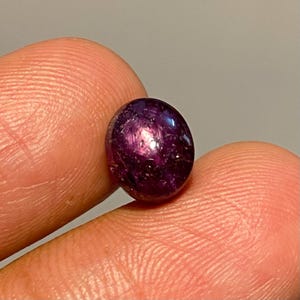 Puede incluir: Una gema redonda y pulida de color púrpura intenso. La piedra tiene una superficie lisa con reflejos internos, creando un efecto de estrella. La gema se sostiene entre dos dedos, mostrando su tamaño y color.