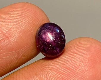 May include: A round, polished gemstone with a deep purple hue. The stone has a smooth surface with internal light reflections, creating a star-like effect. The gem is held between two fingers, showcasing its size and color.