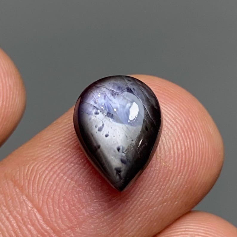 May include: A teardrop-shaped gemstone with a dark, glossy surface. The gem has a deep purple hue with lighter blue and white internal patterns. The stone is held between fingers, showcasing its polished finish and natural beauty.
