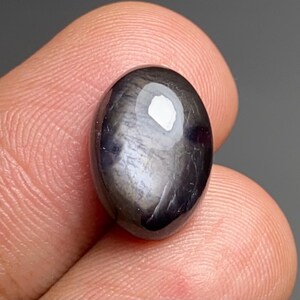 May include: An oval gemstone with a dark, shimmering surface. The stone has a glossy, reflective appearance with a bright white highlight. The gem is held between two fingers, highlighting its size and shape.