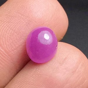 Puede incluir: Una gema magenta vibrante, pulida y de forma ovalada. La gema tiene una superficie lisa y brillante con un reflejo blanco brillante. La piedra se sostiene entre dos dedos, mostrando su color y forma.