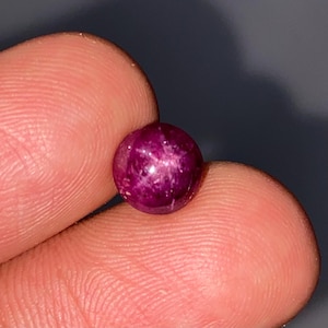 May include: A round, polished, deep magenta gemstone with a star-like effect. The gem is held between two fingers, showcasing its smooth, reflective surface and internal light play. The gem's color is a rich, saturated hue.