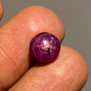 Op de afbeelding: Een ronde, gepolijste edelsteen met een diepe magenta tint, bespikkeld met donkere insluitsels. De edelsteen wordt tussen vingers gehouden, wat de bolvormige vorm en het reflecterende oppervlak benadrukt. De edelsteen is een natuurlijke robijn.