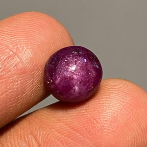 Op de afbeelding: Een ronde, gepolijste edelsteen met een diepe magenta tint. De edelsteen heeft een glad oppervlak met subtiele interne insluitsels en wordt tussen twee vingers gehouden. De edelsteen lijkt een robijn te zijn.