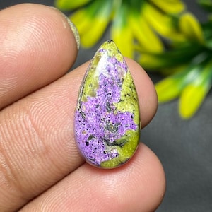 Puede incluir: Una piedra preciosa en forma de lágrima. La piedra presenta una base verde oliva y está adornada con un llamativo patrón de color púrpura y negro. La superficie pulida refleja la luz, resaltando los intrincados detalles de la piedra natural.