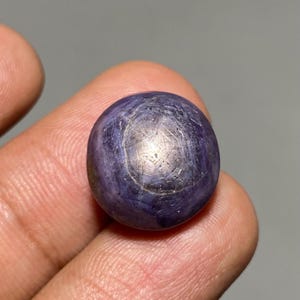 Op de afbeelding: Een close-up van een ronde, gepolijste, donkerblauwe edelsteen. De edelsteen heeft een stervormig patroon op het oppervlak, met een lichtere, zilverachtige kern. De steen wordt tussen vingers gehouden, wat de grootte en vorm laat zien.