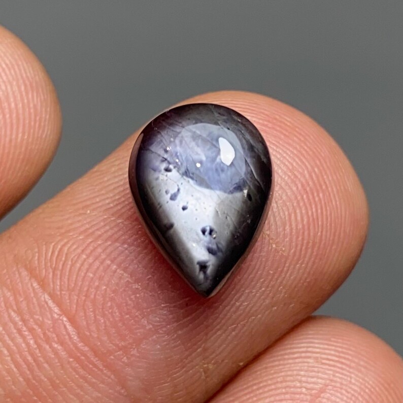 May include: A teardrop-shaped gemstone with a dark, glossy surface. The gem has a deep purple hue with lighter blue and white internal patterns. The stone is held between two fingers against a gray background.