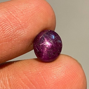 Puede incluir: Una gema pulida, de forma ovalada, de color magenta intenso. La gema tiene un efecto de estrella, con la luz reflejándose en su superficie. La piedra se sostiene entre los dedos, mostrando su color y textura. Esta gema es adecuada para la fabricación de joyas.