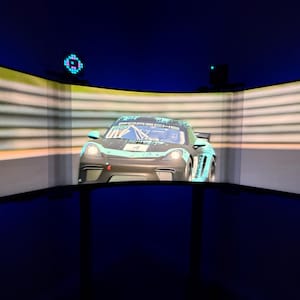 以下が含まれることがあります： 暗いグレーとティールのレーシングカーで、ボンネットに「ポルシェ」の文字が書かれています。湾曲したスクリーンに表示されています。車は水平線のある背景の前にあります。シーンは濃い青色で縁取られています。