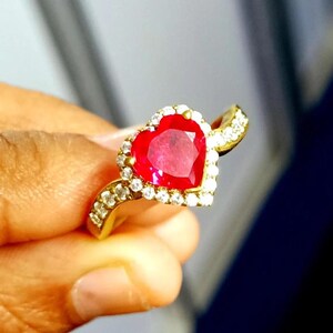 Puede incluir: Un anillo de oro con una piedra preciosa roja brillante en forma de corazón. El corazón está rodeado por un halo de piedras preciosas claras más pequeñas. La banda del anillo también está adornada con piedras preciosas claras.