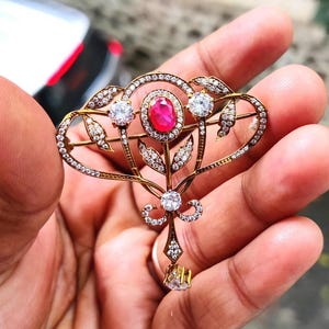 Puede incluir: Broche dorado con una piedra central de rubí ovalada, rodeada de gemas claras y delicados diseños de hojas y flores. El broche tiene una parte superior en forma de corazón y un elemento inferior colgante, que muestra una intrincada artesanía.