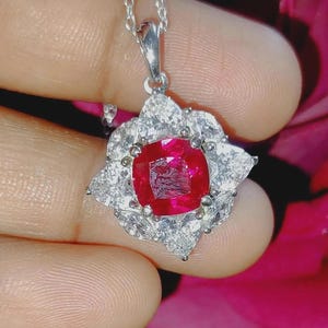 May include: A silver pendant necklace featuring a vibrant red gemstone center, surrounded by clear, sparkling accent stones. The pendant has a floral design, and the chain is delicate.