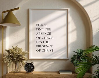 May include: A framed print with the text "PEACE ISN'T THE ABSENCE OF CHAOS IT'S THE PRESENCE OF CHRIST." The print is displayed on a wooden shelf with a clock, a vase of dried flowers, and a small potted plant. A gold lamp is to the left.