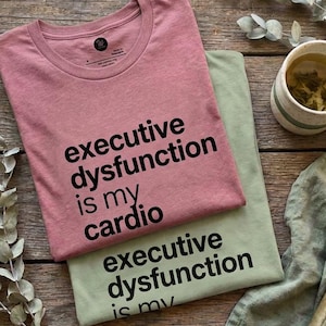 May include: A dusty rose t-shirt and a sage green t-shirt, both featuring the black text "executive dysfunction is my cardio" printed on the front. The shirts are layered together on a wooden surface with a book and a mug.