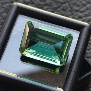 May include: A rectangular, emerald-cut gemstone with a deep teal color is displayed in a black square case. The gem has a faceted surface that reflects light, showcasing its clarity and brilliance. The case is slightly open, revealing a glimpse of the dark surface beneath.