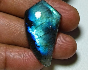 May include: A polished, shield-shaped labradorite gemstone. The stone displays a vibrant play of color, with iridescent blues, greens, and hints of gold. The surface has a smooth, reflective quality, showcasing natural patterns and inclusions. The gemstone is held in a hand.