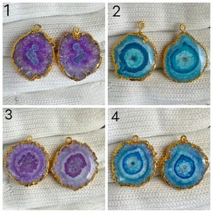 May include: A collection of eight irregular, circular agate slice pendants with gold-toned metal edges and attached loops. The set includes four pairs of pendants in shades of purple and blue, each featuring unique crystalline patterns and textures.