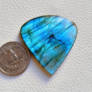 May include: A polished, teardrop-shaped labradorite gemstone, displaying iridescent blue and green hues. The stone is next to a U.S. quarter for size comparison. The gemstone's surface has natural veining and a translucent quality.