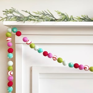 May include: A colorful felt ball garland with red, pink, green, white, and blue balls. The garland is hanging on a white mantel.