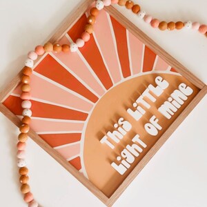 May include: A wooden framed wall art piece with a sunset design in shades of orange, pink, and white. The text "This little light of mine" is written in white letters on the orange background.