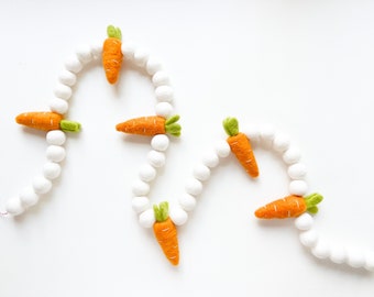 May include: A white felt garland with orange felt carrots. The garland is strung together with white pom poms.
