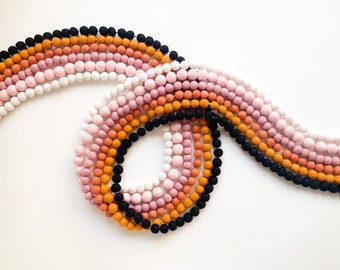 Könnte beinhalten: Eine Girlande aus Filzkugeln in den Farben Schwarz, Orange, Rosa und Weiß. Die Kugeln sind zu einer durchgehenden Schleife zusammengefügt und bilden so eine farbenfrohe und dekorative Dekoration.