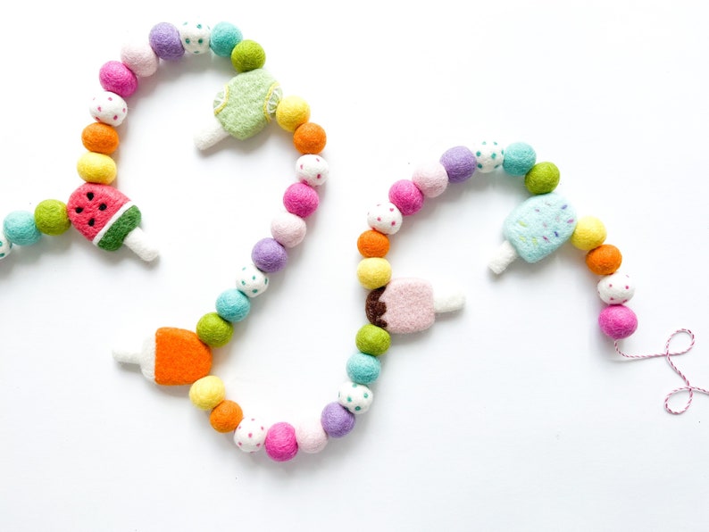 May include: A colorful felt garland with a string of beads and felt popsicle shapes. The popsicles are decorated with watermelon, orange, lime, chocolate, and sprinkles.