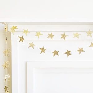 May include: A gold glitter star garland is draped across a white surface. The star-shaped decorations are strung together, creating a festive and decorative element. The garland is ideal for parties and celebrations.
