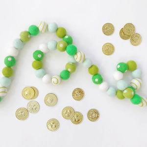 May include: A green and white felt ball garland with shamrock-shaped gold coins scattered around it. The garland is made of felt balls in shades of green and white, with some of the balls having white polka dots. The gold coins are scattered around the garland, and some are overlapping.