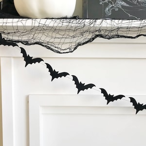 May include: Black bat garland strung across a white surface, with a black decorative net and a white pumpkin in the background. The bats are evenly spaced and add a spooky touch to the decor.