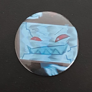 May include: A round button with a cartoon illustration of a blue, blocky face with red eyes and a jagged mouth. The button has a glossy finish and is set against a dark background.