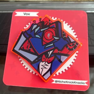 May include: A red, white, blue, and gray Vox pin with a cartoon character design. The pin is on a red card with the name "Vox" and the shop's social media handle, "@NicheKnickKnacks."