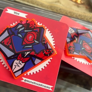 May include: Two enamel pins on red cards. The pins feature a stylized character design in blue, red, and white with an orange border. The cards have the name "Vox" and the seller's social media handle. The pins are on a reflective surface.
