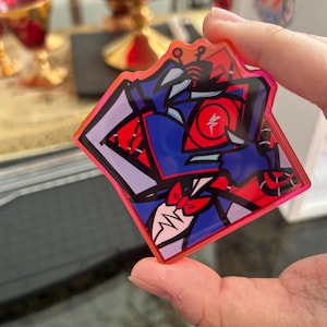 May include: A square-shaped, colorful sticker with a cartoon character design. The sticker features a red, blue, and purple color scheme with a pink border. The character has a red bow tie and a lightning bolt symbol.