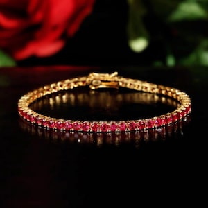 Könnte beinhalten: Ein goldfarbenes Tennisarmband mit einer durchgehenden Reihe runder, leuchtend roter Edelsteine. Das Armband ist mit einem oben sichtbaren Metallverschluss gesichert und zeigt ein einheitliches Design mit polierten Metallfassungen.