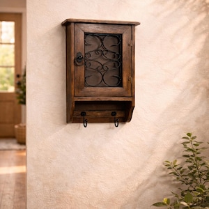 Op de afbeelding: Een donkerhouten wandkast met een glazen deur en decoratief metalen krulwerk. Onder de kast bevinden zich twee haken. De kast is bevestigd aan een getextureerde beige muur.