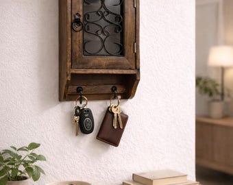 Op de afbeelding: Een aan de muur gemonteerde houten sleutelkast met een donkere afwerking en een decoratief smeedijzeren ontwerp op de glazen deur. Onder de kast bevinden zich sleutelhaken met sleutels en een leren sleutelhouder. Een kleine plant en een kom met sleutels staan op een houten tafel.