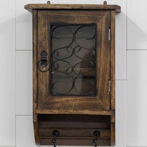 Op de afbeelding: Een rustieke houten sleutelkast met een donkerbruine afwerking. De kast heeft een glazen deur met een decoratief zwart metalen krulontwerp. Onder de deur zitten twee sleutelhaken. De kast is aan een witte muur bevestigd.