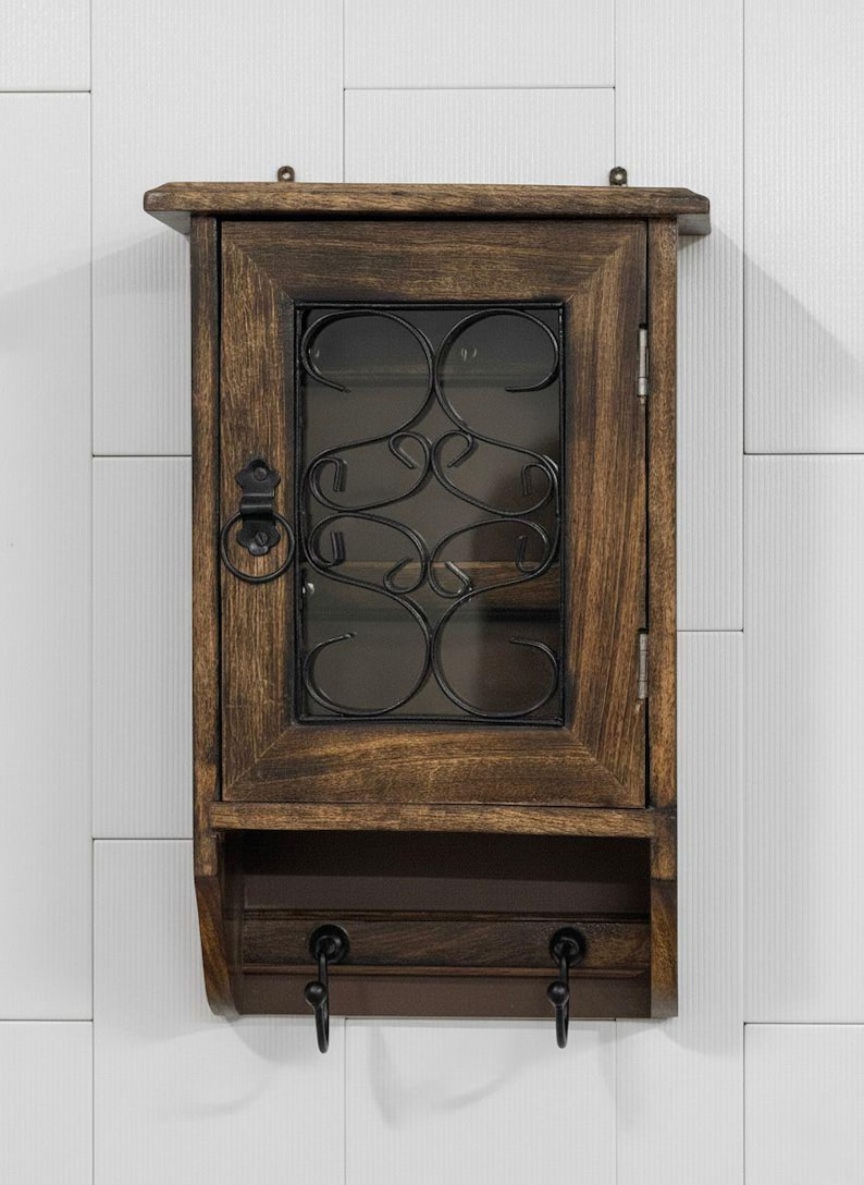 Op de afbeelding: Een rustieke houten sleutelkast met een donkerbruine afwerking. De kast heeft een glazen deur met een decoratief zwart metalen krulontwerp. Onder de deur zitten twee sleutelhaken. De kast is aan een witte muur bevestigd.
