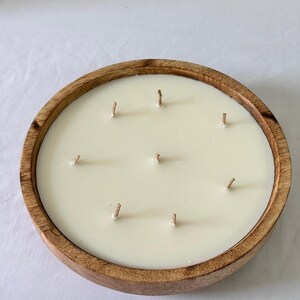 May include: A large, round candle with eight wicks, set in a natural wood bowl. The candle wax is a creamy off-white color. The wooden bowl has a warm, light brown tone, creating a rustic aesthetic. The candle is designed for home fragrance.