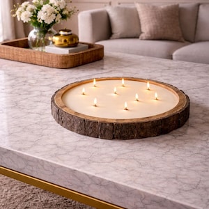 May include: A large, circular candle with several lit wicks, set in a wooden holder resembling a tree trunk. The candle rests on a marble coffee table. A tray with flowers and a sofa are visible in the background.