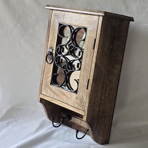 Op de afbeelding: Een houten wandkast met een donkere afwerking. De kast heeft een decoratief metalen ontwerp achter de glazen deur en twee zwarte haken eronder. Ontworpen voor opslag en organisatie.
